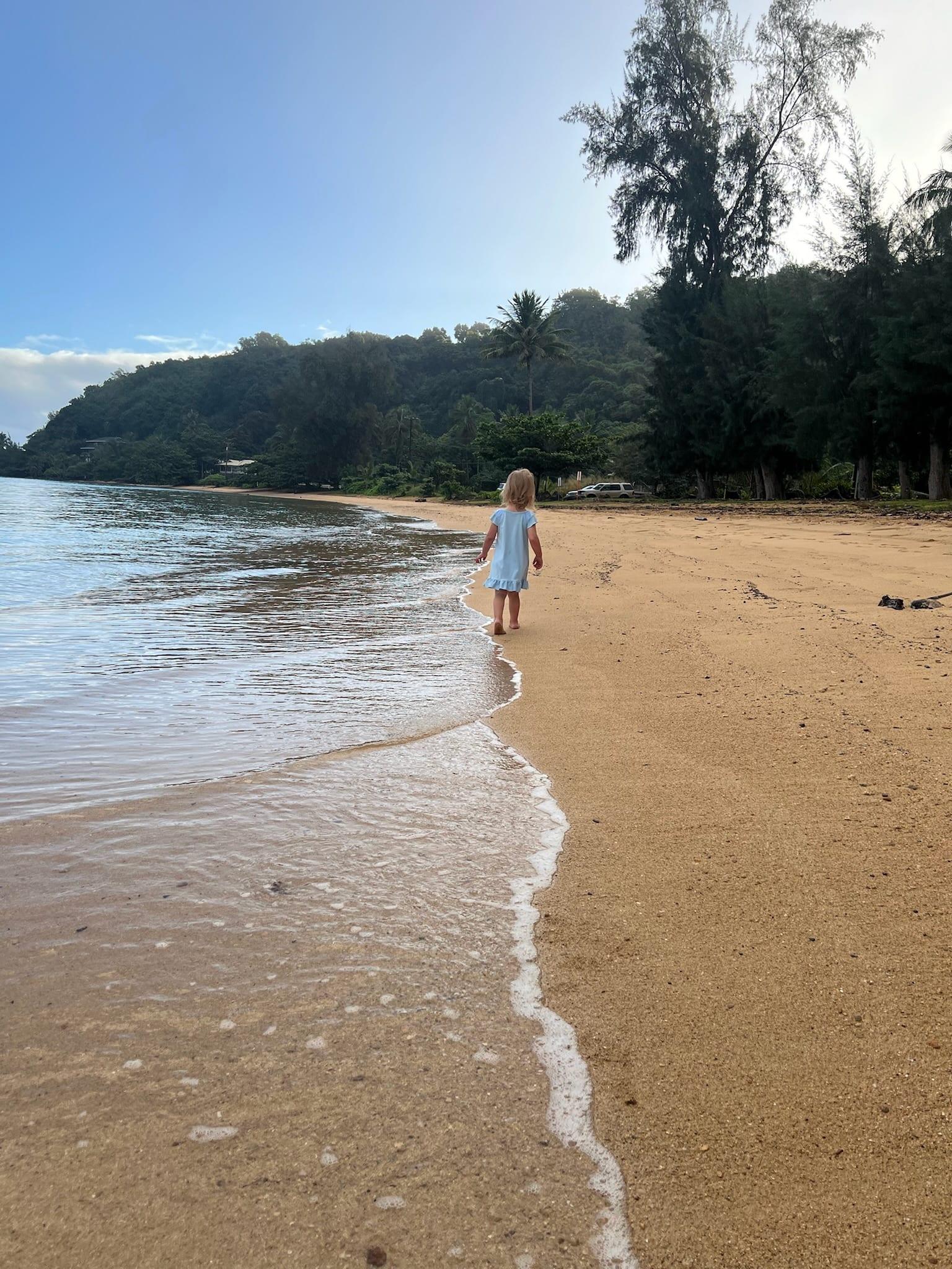 A young child walks the calm, glassy shoreline at Anini Beach, the gentlest water on Kauai's North Shore