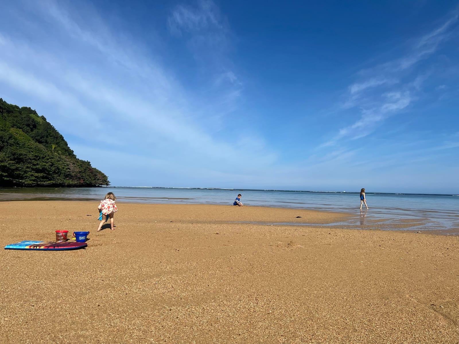Kids playing on a calm, sunny North Shore beach with the reef visible offshore, a typical beach day from Princeville