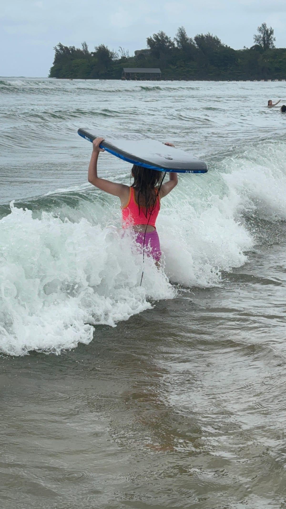 Girl carrying a boogie board through the water at Hanalei Bay, a classic North Shore Kauai beach day with kids