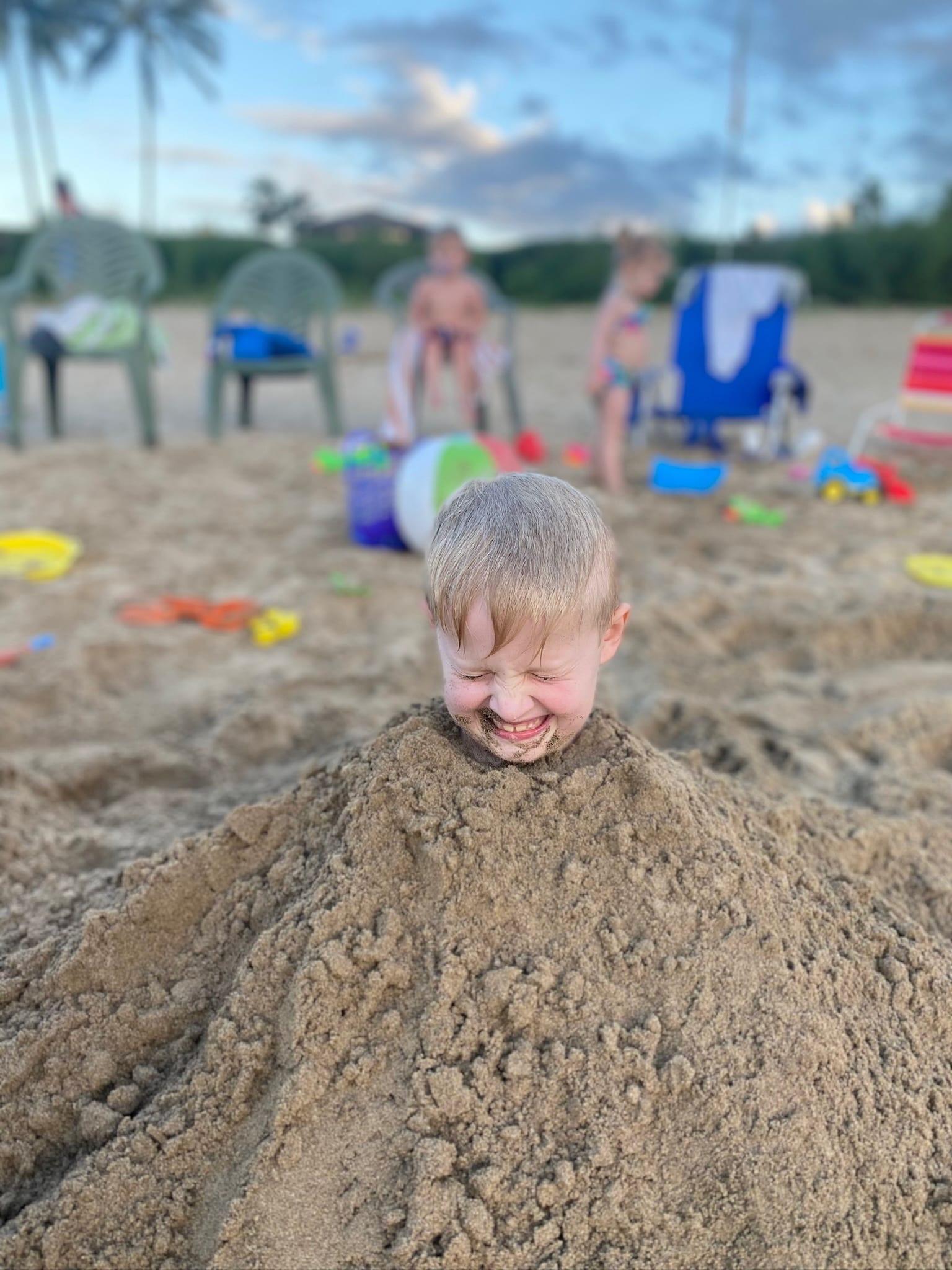 A grinning boy buried up to his neck in sand at golden hour, with other kids and beach chairs behind
