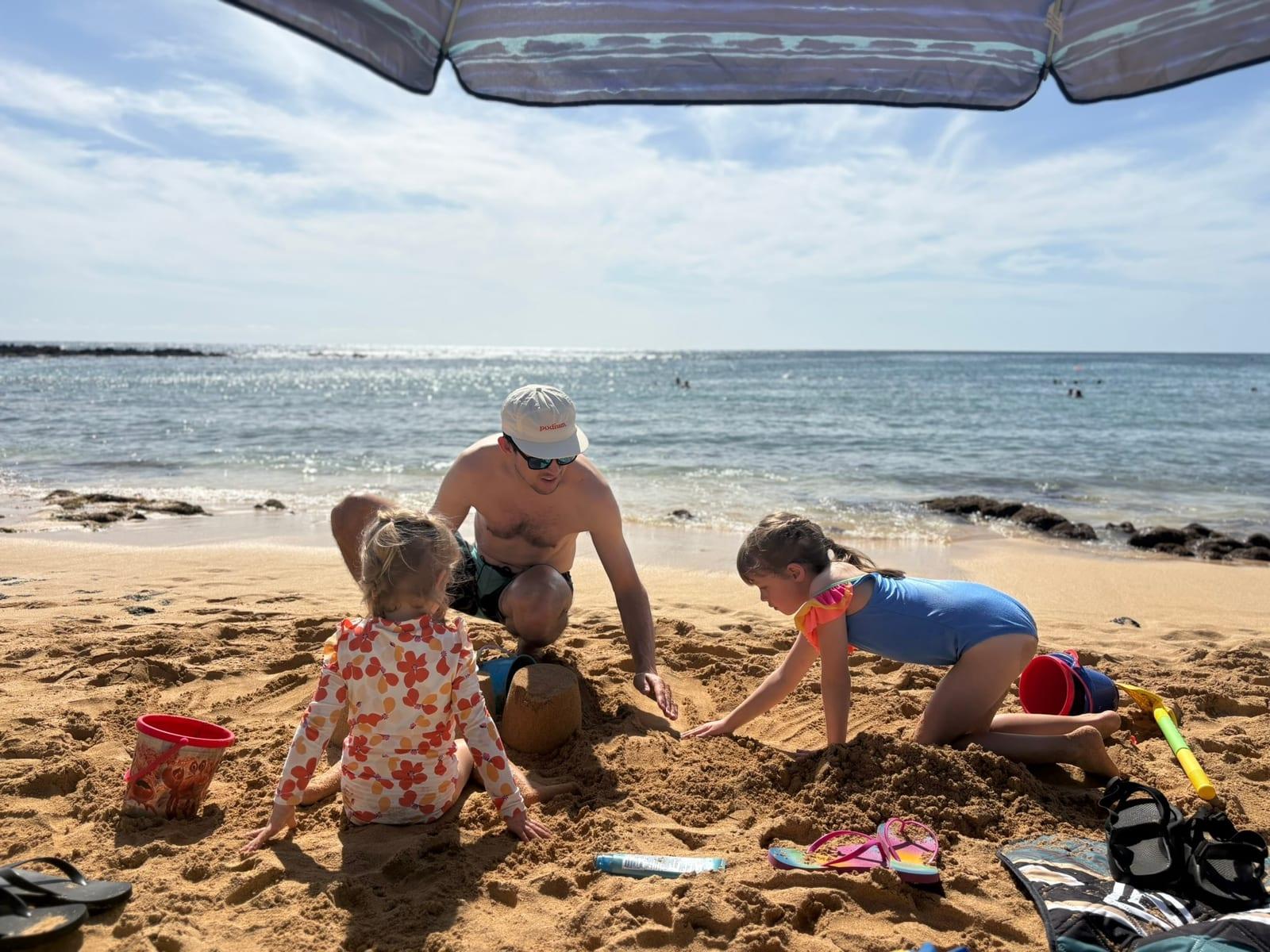 Three kids and a dad building a sandcastle under a beach umbrella on a sunny Kauai beach