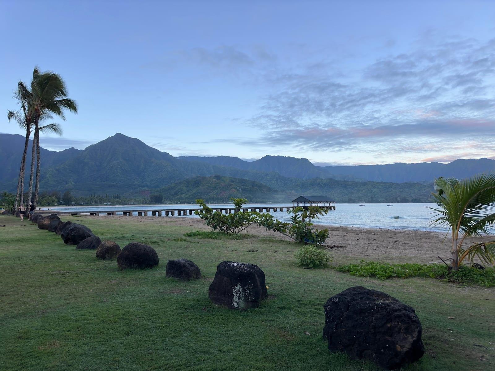 Hanalei Bay at dawn with lava-rock boulders on the lawn, pier, and green mountains near Princeville