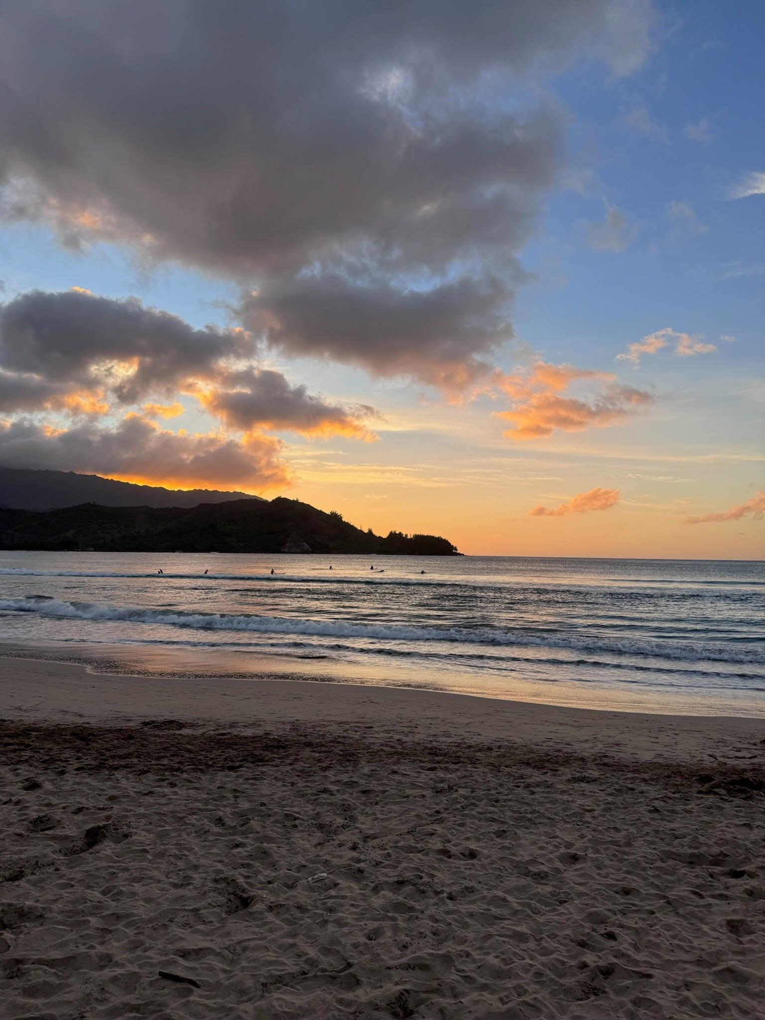 Surfers in the water at Hanalei Bay as the sun sets behind the North Shore mountains